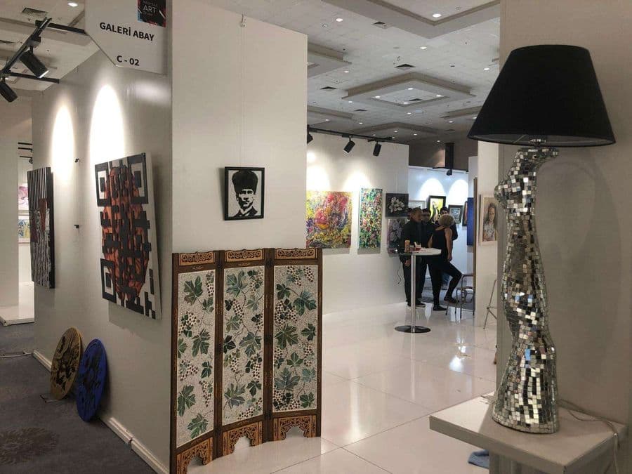 Sanat fuarında "Galeri Abay C-02" standının girişi. Stantta ahşap bir paravan ve çeşitli tablolar yer alırken, ön planda mozaik aynalı modern bir lambader duruyor.