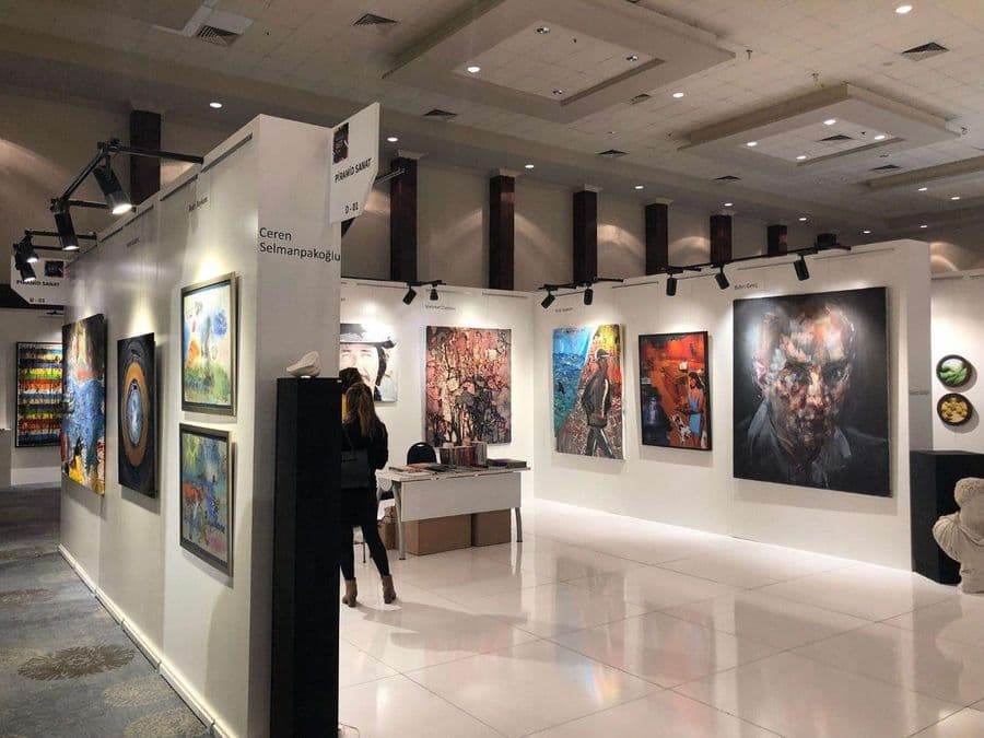Sanat fuarında yer alan "Piramid Sanat D-02" standının içten görünümü. Parlak beyaz zeminli alanda, duvarlarda büyük bir portre, soyut tablolar ve farklı eserler spot ışıkları altında sergileniyor.