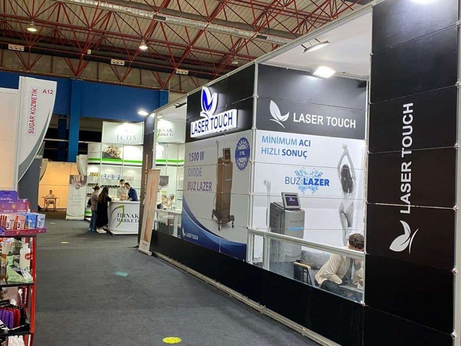 "Laser Touch" markasına ait, estetik ve lazer cihazlarının sergilendiği, kapalı bir ticaret fuarı standı. Standın sağ tarafında "2500W Yeni Nesil IPL" yazılı büyük bir tabela ve bir cihaz sergileniyor. Beyaz raflarda kozmetik ürünler, stant zemininde ise "ICEBERG" yazılı bir cihaz da dahil olmak üzere estetik cihazlar yer alıyor.