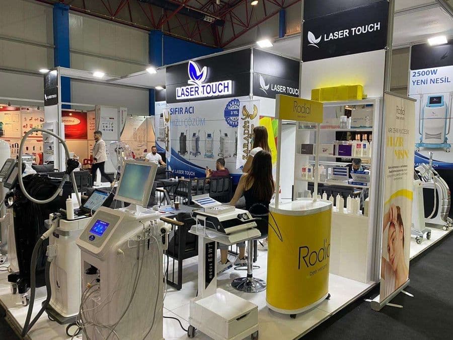 "Laser Touch" markasına ait, estetik ve lazer cihazlarının sergilendiği, kapalı bir ticaret fuarı standı. Standın sağ tarafında "2500W Yeni Nesil IPL" yazılı büyük bir tabela ve bir cihaz sergileniyor. Beyaz raflarda kozmetik ürünler, stant zemininde ise "ICEBERG" yazılı bir cihaz da dahil olmak üzere estetik cihazlar yer alıyor.