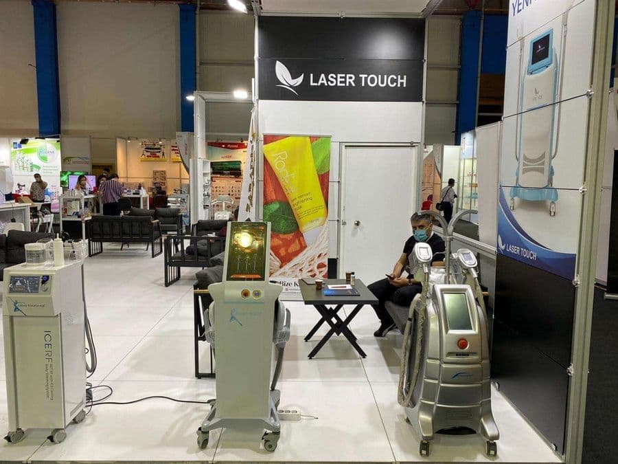 "Laser Touch" markasına ait, estetik ve lazer cihazlarının sergilendiği, kapalı bir ticaret fuarı standı. Standın sağ tarafında "2500W Yeni Nesil IPL" yazılı büyük bir tabela ve bir cihaz sergileniyor. Beyaz raflarda kozmetik ürünler, stant zemininde ise "ICEBERG" yazılı bir cihaz da dahil olmak üzere estetik cihazlar yer alıyor.
