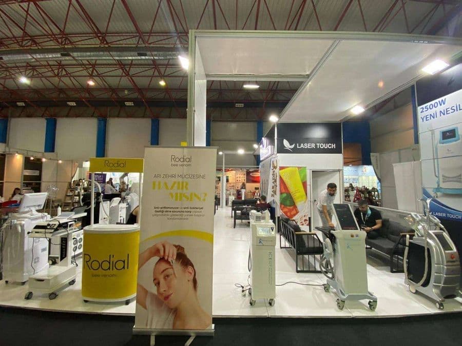 "Laser Touch" markasına ait, estetik ve lazer cihazlarının sergilendiği, kapalı bir ticaret fuarı standı. Standın sağ tarafında "2500W Yeni Nesil IPL" yazılı büyük bir tabela ve bir cihaz sergileniyor. Beyaz raflarda kozmetik ürünler, stant zemininde ise "ICEBERG" yazılı bir cihaz da dahil olmak üzere estetik cihazlar yer alıyor.