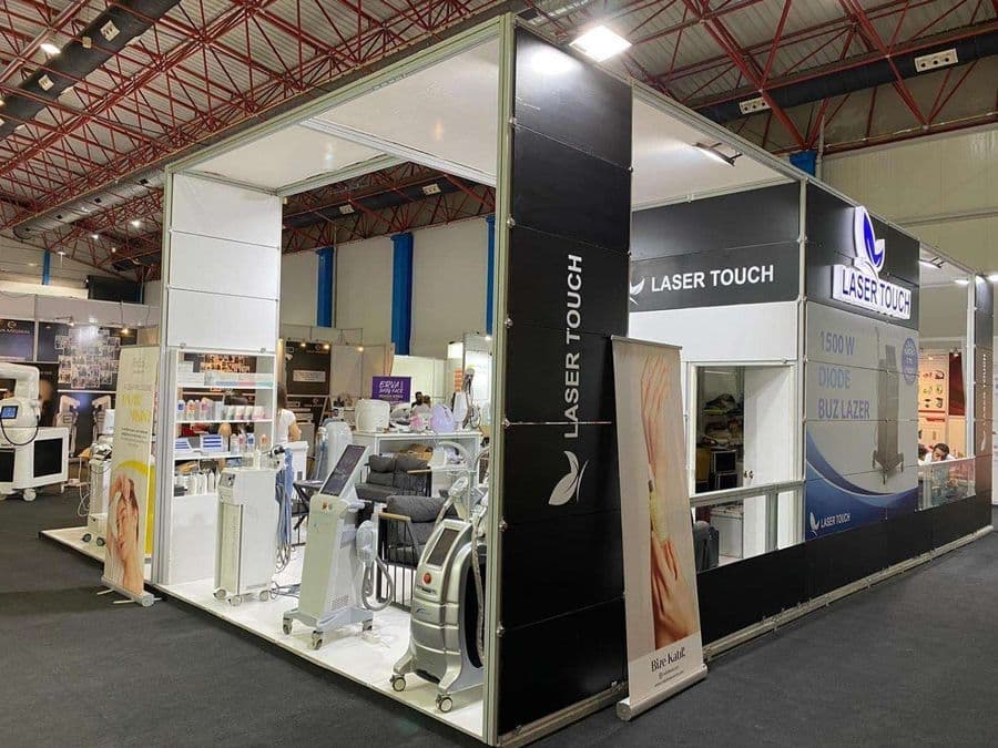 "Laser Touch" markasına ait, estetik ve lazer cihazlarının sergilendiği, kapalı bir ticaret fuarı standı. Standın sağ tarafında "2500W Yeni Nesil IPL" yazılı büyük bir tabela ve bir cihaz sergileniyor. Beyaz raflarda kozmetik ürünler, stant zemininde ise "ICEBERG" yazılı bir cihaz da dahil olmak üzere estetik cihazlar yer alıyor.