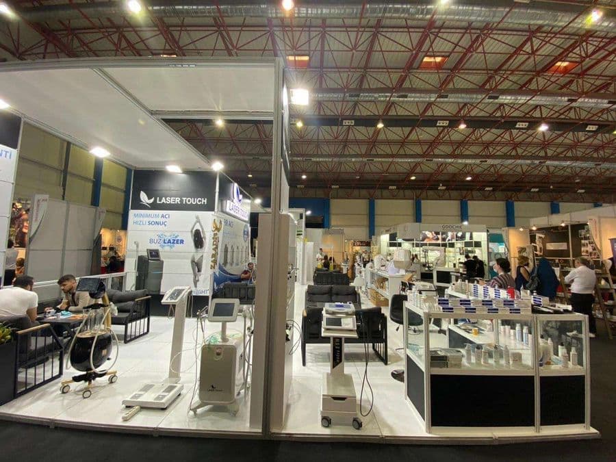 "Laser Touch" markasına ait, estetik ve lazer cihazlarının sergilendiği, kapalı bir ticaret fuarı standı. Standın sağ tarafında "2500W Yeni Nesil IPL" yazılı büyük bir tabela ve bir cihaz sergileniyor. Beyaz raflarda kozmetik ürünler, stant zemininde ise "ICEBERG" yazılı bir cihaz da dahil olmak üzere estetik cihazlar yer alıyor.