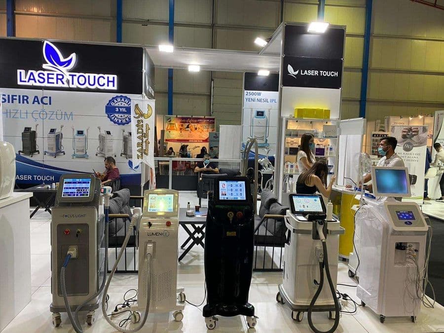 "Laser Touch" markasına ait, estetik ve lazer cihazlarının sergilendiği, kapalı bir ticaret fuarı standı. Standın sağ tarafında "2500W Yeni Nesil IPL" yazılı büyük bir tabela ve bir cihaz sergileniyor. Beyaz raflarda kozmetik ürünler, stant zemininde ise "ICEBERG" yazılı bir cihaz da dahil olmak üzere estetik cihazlar yer alıyor.