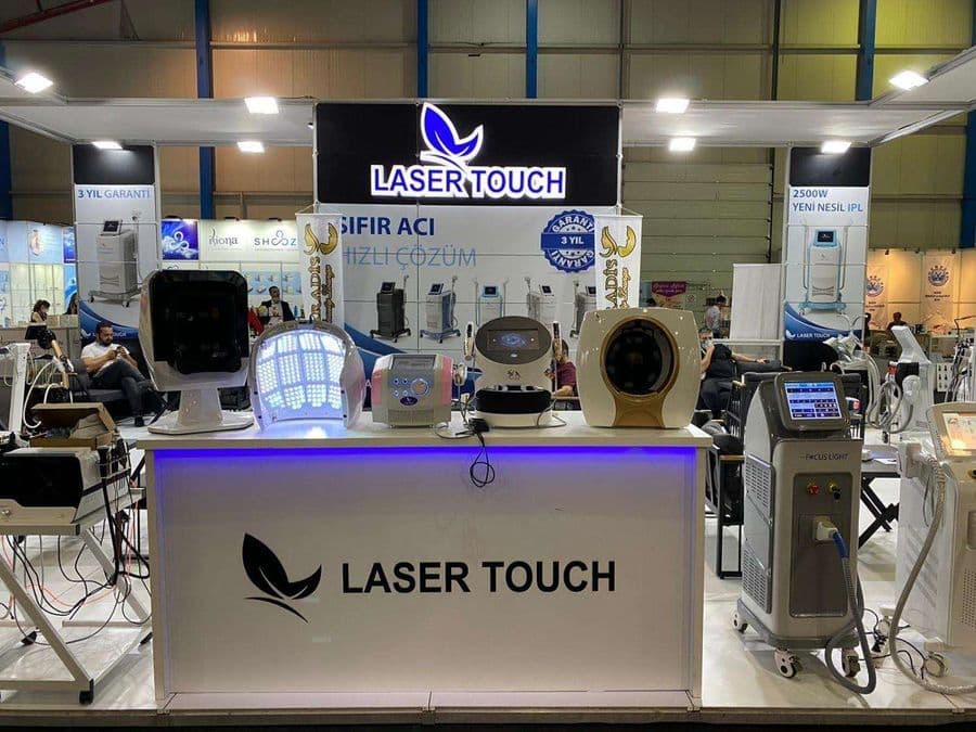 "Laser Touch" markasına ait, estetik ve lazer cihazlarının sergilendiği, kapalı bir ticaret fuarı standı. Standın sağ tarafında "2500W Yeni Nesil IPL" yazılı büyük bir tabela ve bir cihaz sergileniyor. Beyaz raflarda kozmetik ürünler, stant zemininde ise "ICEBERG" yazılı bir cihaz da dahil olmak üzere estetik cihazlar yer alıyor.