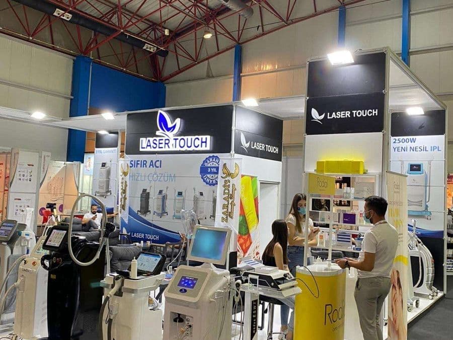 "Laser Touch" markasına ait, estetik ve lazer cihazlarının sergilendiği, kapalı bir ticaret fuarı standı. Standın sağ tarafında "2500W Yeni Nesil IPL" yazılı büyük bir tabela ve bir cihaz sergileniyor. Beyaz raflarda kozmetik ürünler, stant zemininde ise "ICEBERG" yazılı bir cihaz da dahil olmak üzere estetik cihazlar yer alıyor.