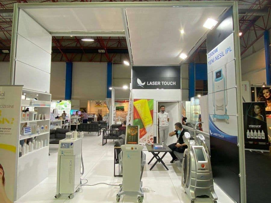 "Laser Touch" markasına ait, estetik ve lazer cihazlarının sergilendiği, kapalı bir ticaret fuarı standı. Standın sağ tarafında "2500W Yeni Nesil IPL" yazılı büyük bir tabela ve bir cihaz sergileniyor. Beyaz raflarda kozmetik ürünler, stant zemininde ise "ICEBERG" yazılı bir cihaz da dahil olmak üzere estetik cihazlar yer alıyor.