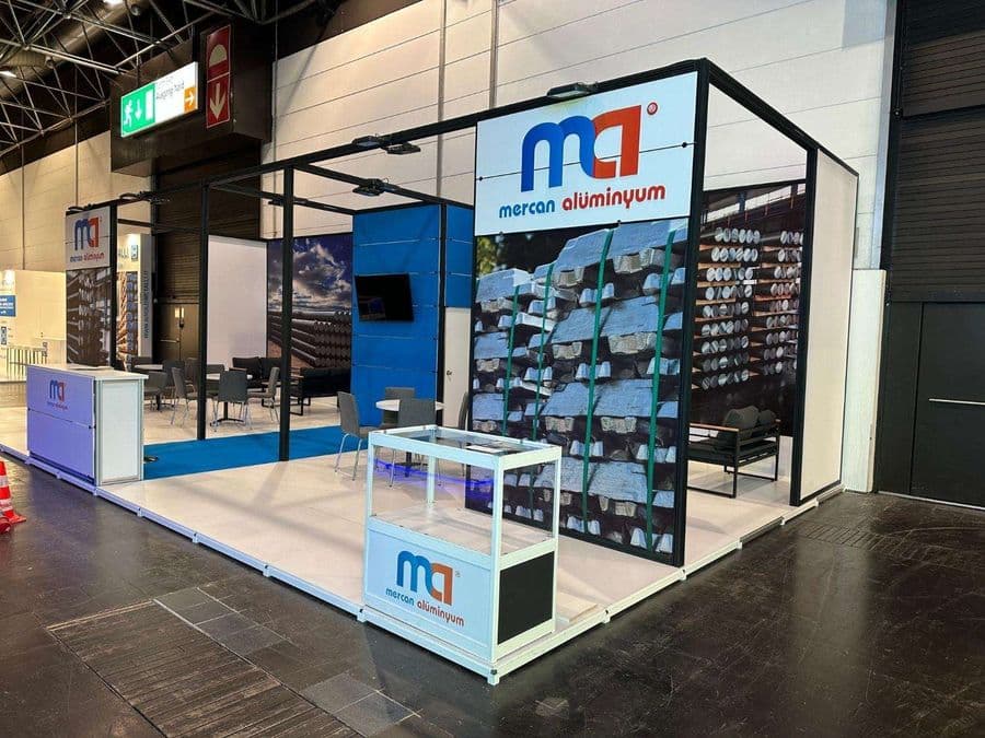 Kapalı bir fuar alanında kurulmuş, "mG Merçan Alüminyum" markasına ait profesyonel bir ticaret fuarı standı. Standın ön yüzünde paletlenmiş alüminyum külçelerinin büyük bir görseli yer alıyor.