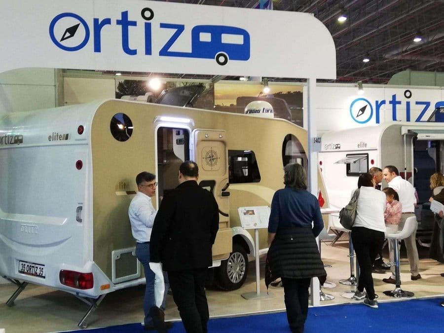 Bir karavan fuarı veya ticaret fuarı standında sergilenen, beyaz renkli, "Ortiz" markalı bir çekme karavan (römork karavan). Mavi halı kaplı alanda karavanın etrafında yürüyen ve inceleyen ziyaretçiler bulunuyor.