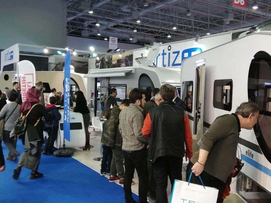 Bir karavan fuarı veya ticaret fuarı standında sergilenen, beyaz renkli, "Ortiz" markalı bir çekme karavan (römork karavan). Mavi halı kaplı alanda karavanın etrafında yürüyen ve inceleyen ziyaretçiler bulunuyor.