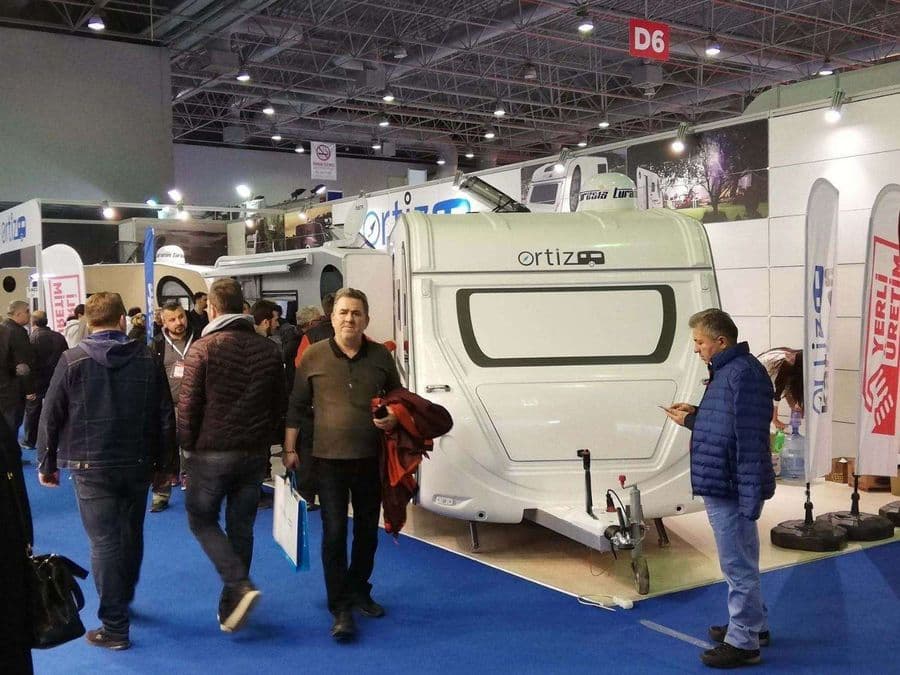 Bir karavan fuarı veya ticaret fuarı standında sergilenen, beyaz renkli, "Ortiz" markalı bir çekme karavan (römork karavan). Mavi halı kaplı alanda karavanın etrafında yürüyen ve inceleyen ziyaretçiler bulunuyor.
