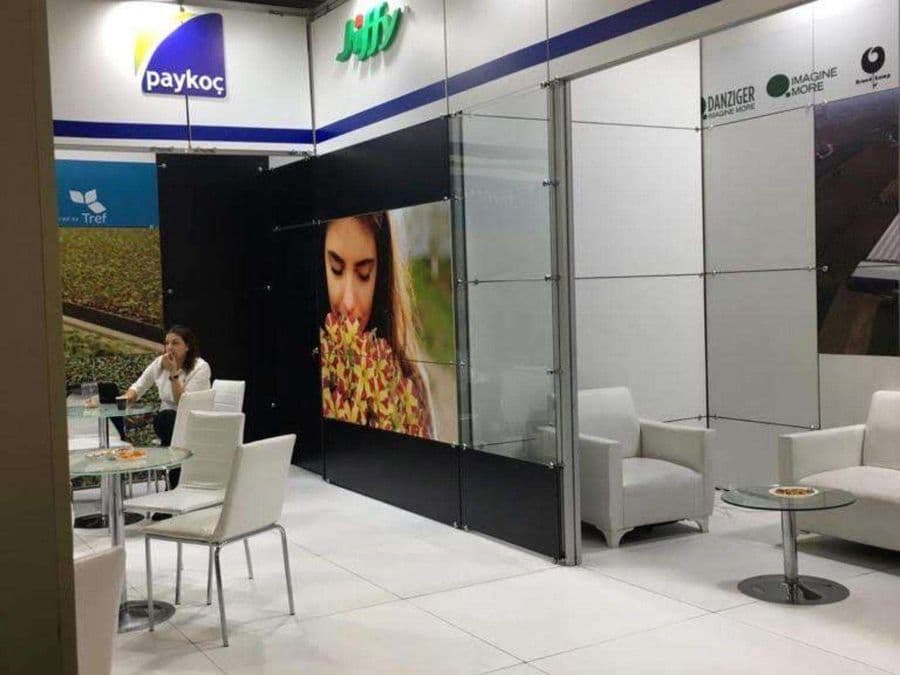 Bahçecilik ürünleri markası "Jiffy"nin bir fuardaki tanıtım standı. Beyaz ve yeşil temalı stantta, bir yanda filizlerin sergilendiği raflar, diğer yanda ise ziyaretçilerin oturduğu masa ve sandalyeler bulunuyor.