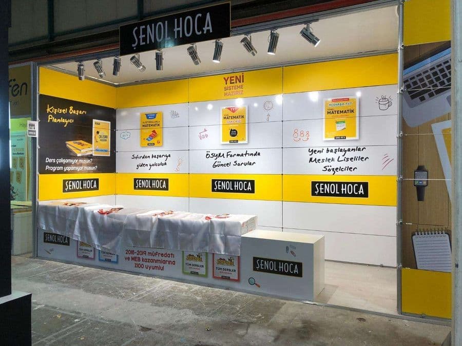 "Şenol Hoca" eğitim markasının bir fuardaki kitap satış standı. Sarı ve beyaz renklerin hakim olduğu stantta, matematik kitapları sergileniyor ve duvarlarda öğrencilere yönelik sloganlar yer alıyor.