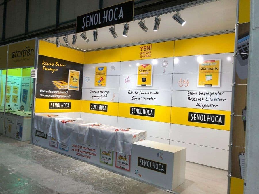 "Şenol Hoca" eğitim markasının bir fuardaki kitap satış standı. Sarı ve beyaz renklerin hakim olduğu stantta, matematik kitapları sergileniyor ve duvarlarda öğrencilere yönelik sloganlar yer alıyor.
