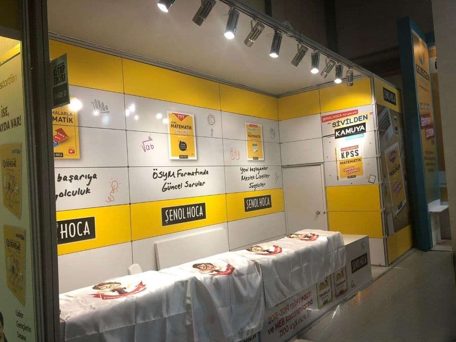 "Şenol Hoca" eğitim markasının bir fuardaki kitap satış standı. Sarı ve beyaz renklerin hakim olduğu stantta, matematik kitapları sergileniyor ve duvarlarda öğrencilere yönelik sloganlar yer alıyor.