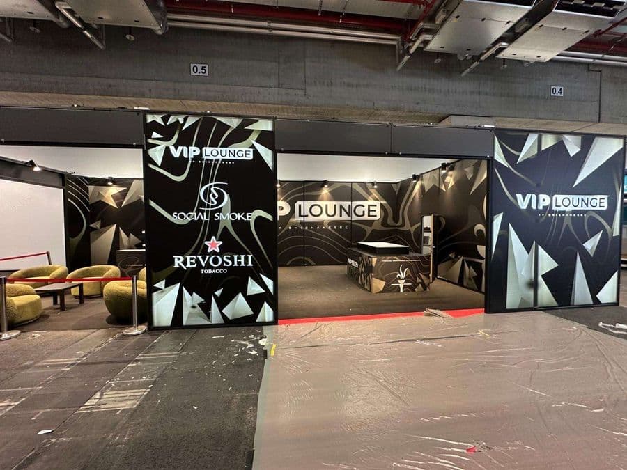 "Revoshi" ve "Social Smoke" markalarının sponsor olduğu bir VIP Lounge'un fuar alanındaki kurulum anı. Siyah ve gold geometrik desenlerle süslü standın önündeki zemin plastik bir örtüyle kaplıdır.