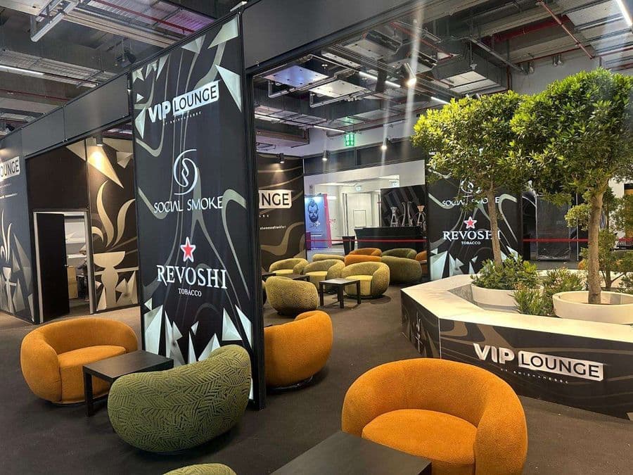 Bir fuar veya kongre merkezinde, kırmızı şeritlerle ayrılmış "VIP Lounge" dinlenme bölümü. Alanda, turuncu ve yeşil renklerde çok sayıda yuvarlak hatlı koltuk ve küçük sehpalar bulunuyor.