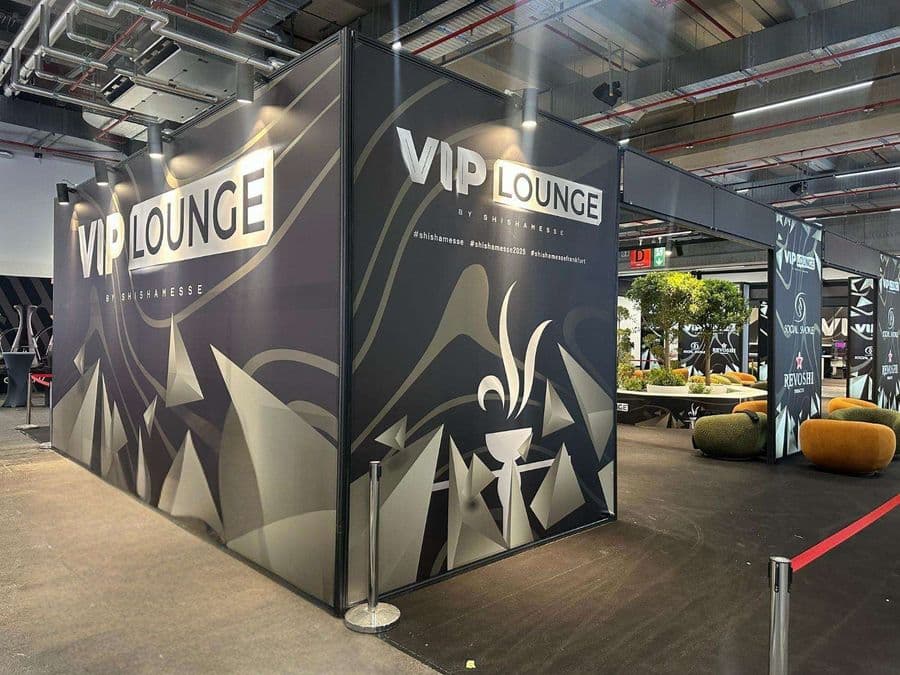 Bir fuar veya kongre merkezinde, kırmızı şeritlerle ayrılmış "VIP Lounge" dinlenme bölümü. Alanda, turuncu ve yeşil renklerde çok sayıda yuvarlak hatlı koltuk ve küçük sehpalar bulunuyor.