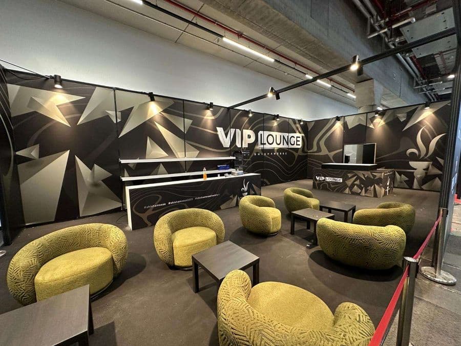 Bir fuar veya kongre merkezinde, kırmızı şeritlerle ayrılmış "VIP Lounge" dinlenme bölümü. Alanda, turuncu ve yeşil renklerde çok sayıda yuvarlak hatlı koltuk ve küçük sehpalar bulunuyor.