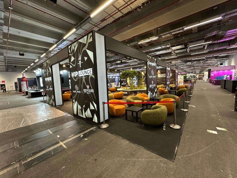 Bir fuar veya kongre merkezinde, kırmızı şeritlerle ayrılmış "VIP Lounge" dinlenme bölümü. Alanda, turuncu ve yeşil renklerde çok sayıda yuvarlak hatlı koltuk ve küçük sehpalar bulunuyor.