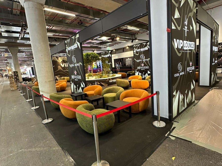 Bir fuar veya kongre merkezinde, kırmızı şeritlerle ayrılmış "VIP Lounge" dinlenme bölümü. Alanda, turuncu ve yeşil renklerde çok sayıda yuvarlak hatlı koltuk ve küçük sehpalar bulunuyor.