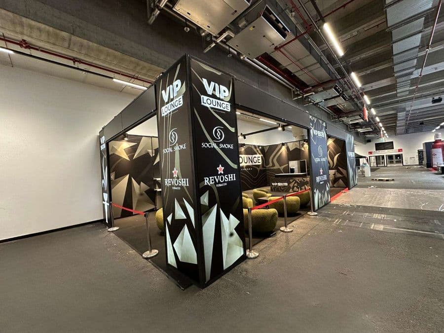 Bir fuar veya kongre merkezinde, kırmızı şeritlerle ayrılmış "VIP Lounge" dinlenme bölümü. Alanda, turuncu ve yeşil renklerde çok sayıda yuvarlak hatlı koltuk ve küçük sehpalar bulunuyor.