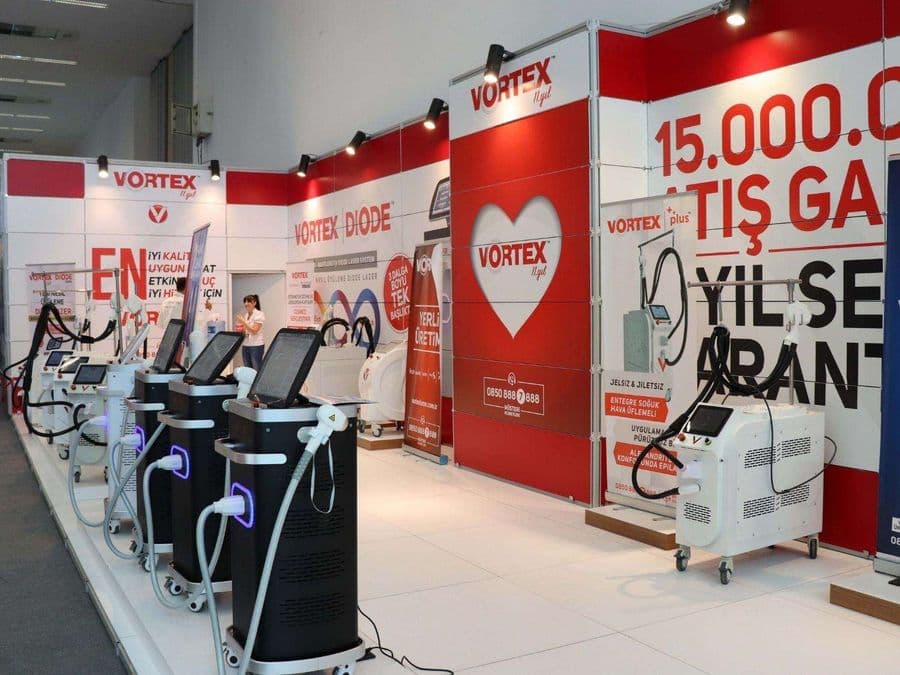 "Vortex Diode Lazer" markasının medikal estetik cihazlarını sergilediği bir fuar standı. Kırmızı ve beyaz temalı stantta, çok sayıda lazer cihazı yan yana dizilerek tanıtılıyor.
