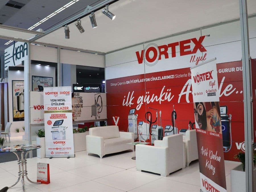 "Vortex Diode Lazer" markasının medikal estetik cihazlarını sergilediği bir fuar standı. Kırmızı ve beyaz temalı stantta, çok sayıda lazer cihazı yan yana dizilerek tanıtılıyor.