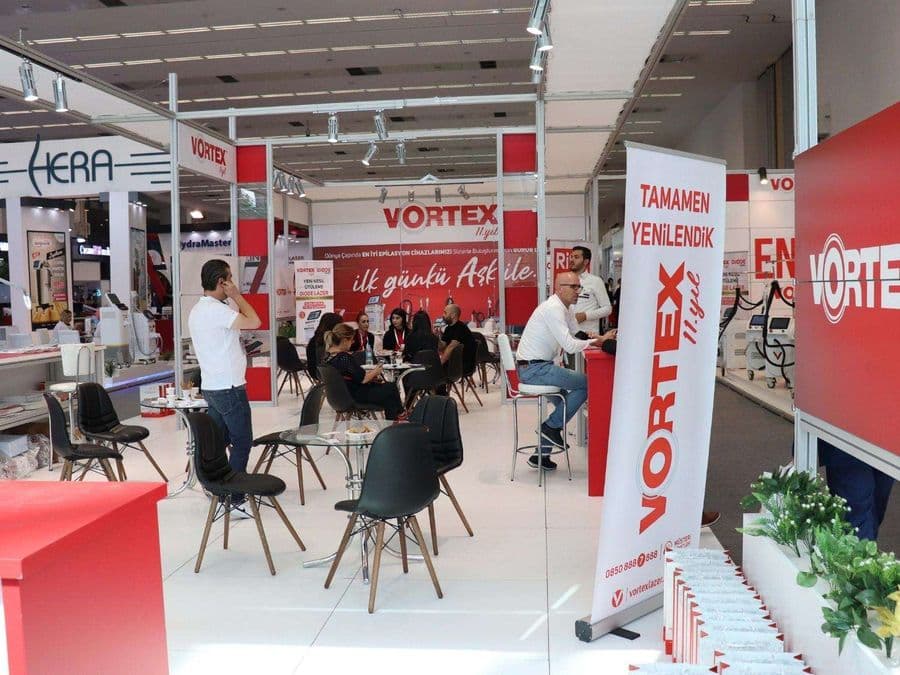 Endüstriyel ürünler fuarında, "VORTEX" markasının modern standı. Işıklı büyük bir logoya sahip standın içi, ziyaretçiler için çok sayıda kanepe ve koltukla bir bekleme alanı gibi tasarlanmış.