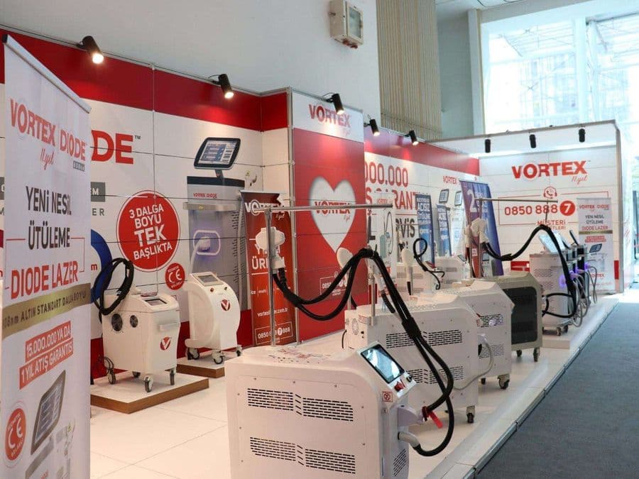 "Vortex Diode Lazer" markasının medikal estetik cihazlarını sergilediği bir fuar standı. Kırmızı ve beyaz temalı stantta, çok sayıda lazer cihazı yan yana dizilerek tanıtılıyor.