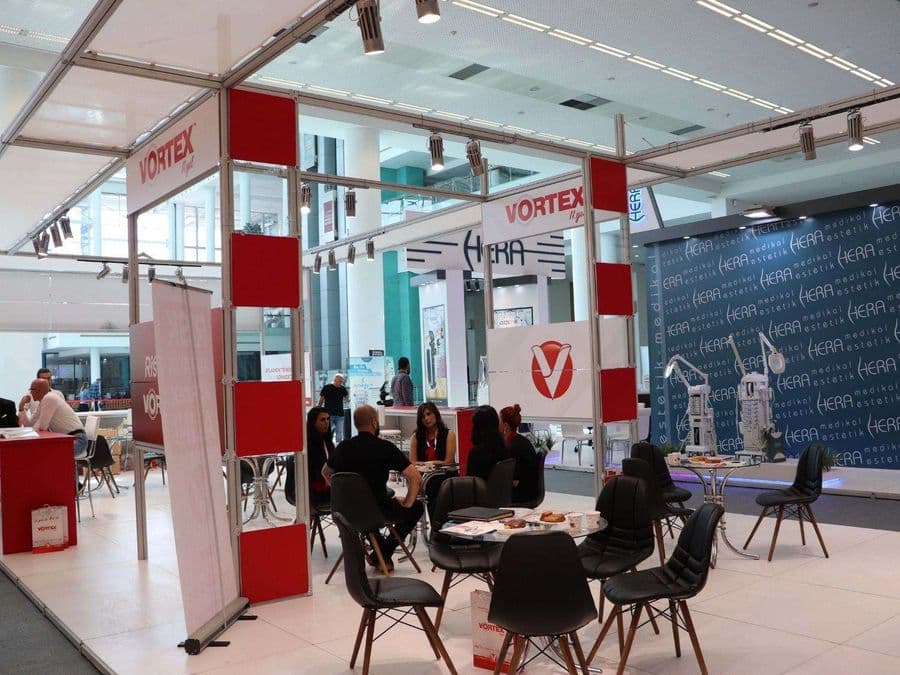 Endüstriyel ürünler fuarında, "VORTEX" markasının modern standı. Işıklı büyük bir logoya sahip standın içi, ziyaretçiler için çok sayıda kanepe ve koltukla bir bekleme alanı gibi tasarlanmış.
