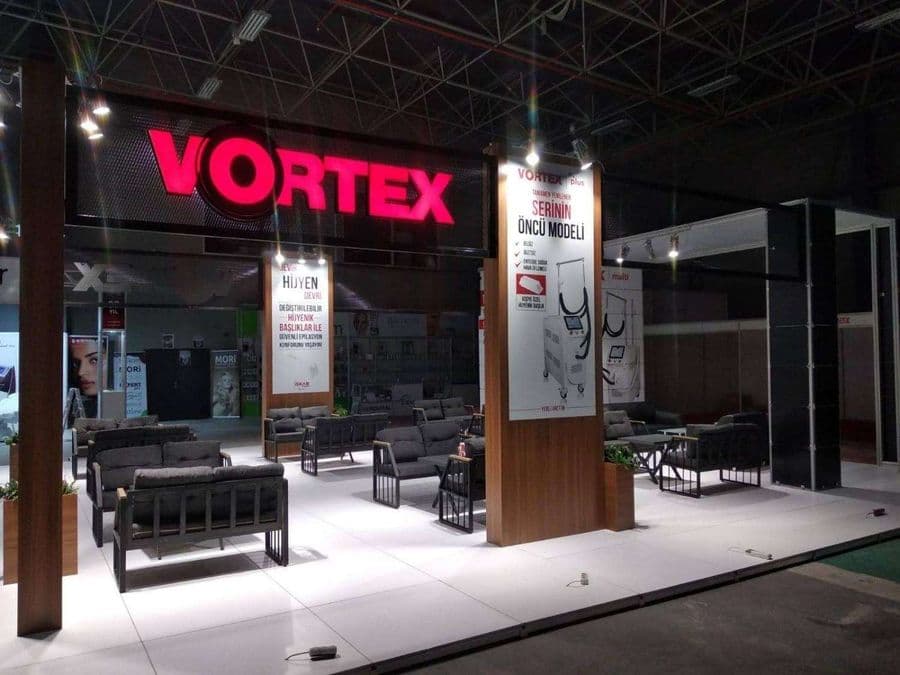 Endüstriyel ürünler fuarında, "VORTEX" markasının modern standı. Işıklı büyük bir logoya sahip standın içi, ziyaretçiler için çok sayıda kanepe ve koltukla bir bekleme alanı gibi tasarlanmış.