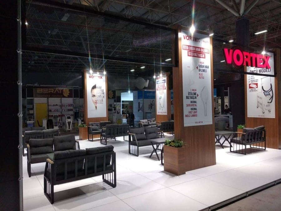 Endüstriyel ürünler fuarında, "VORTEX" markasının modern standı. Işıklı büyük bir logoya sahip standın içi, ziyaretçiler için çok sayıda kanepe ve koltukla bir bekleme alanı gibi tasarlanmış.